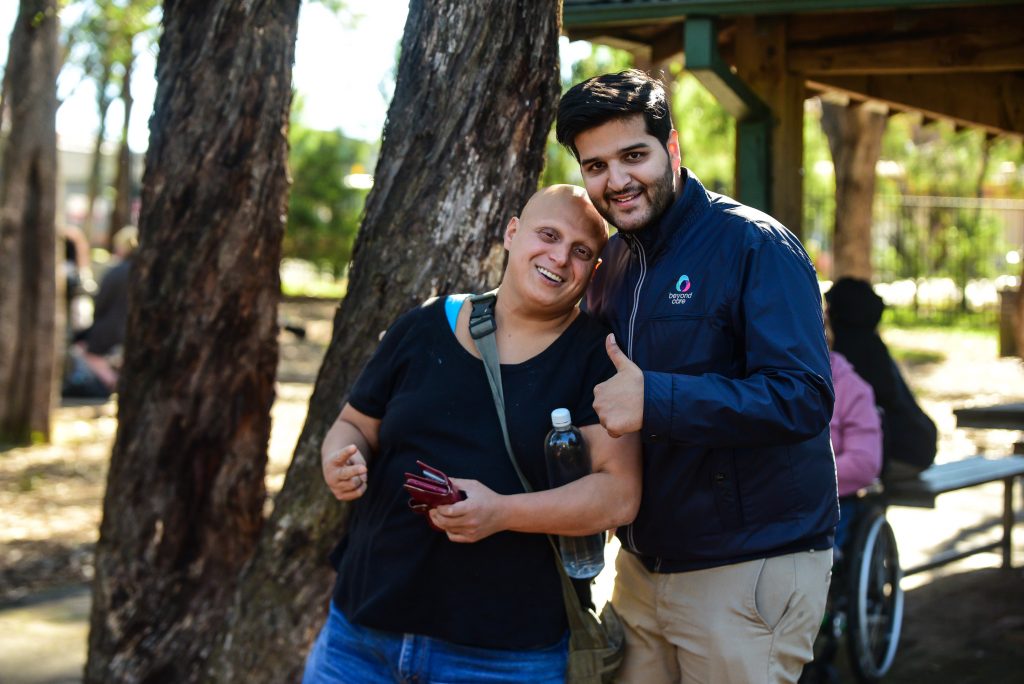 Two people are outdoors near trees. One person is smiling and holding keys; the other is giving a thumbs-up and holding a water bottle. A wheelchair is visible in the background.