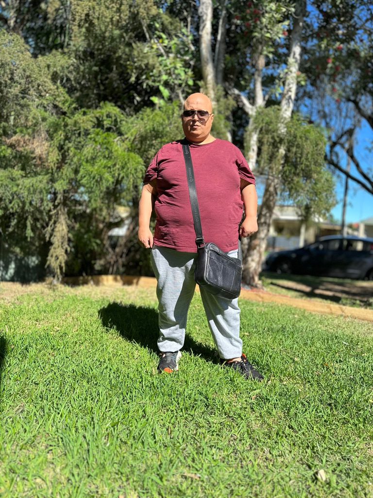 A person stands on the grass in front of their SIL accommodation, wearing a maroon shirt, gray pants, black shoes, and sunglasses, with a black crossbody bag. Trees and a parked car provide a serene background to this Sydney scene.