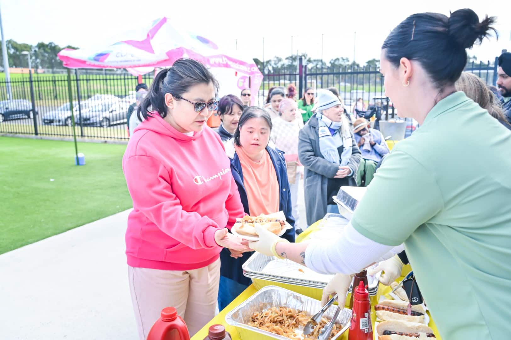 At an outdoor event featuring a spread of hot dogs and condiments, people are warmly served. A person in a pink hoodie receives a plate from the server, reminiscent of the personalized attention seen in NDIS home care settings.