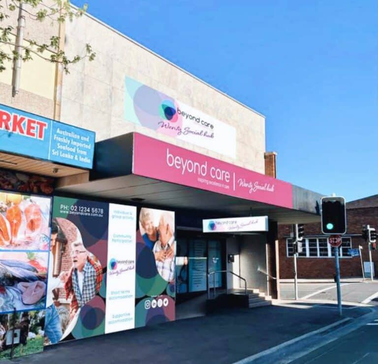 A building facade with a pink sign reading "Beyond Care" and "Nursing & Specialist Health." A contact number and various images are displayed on the storefront.