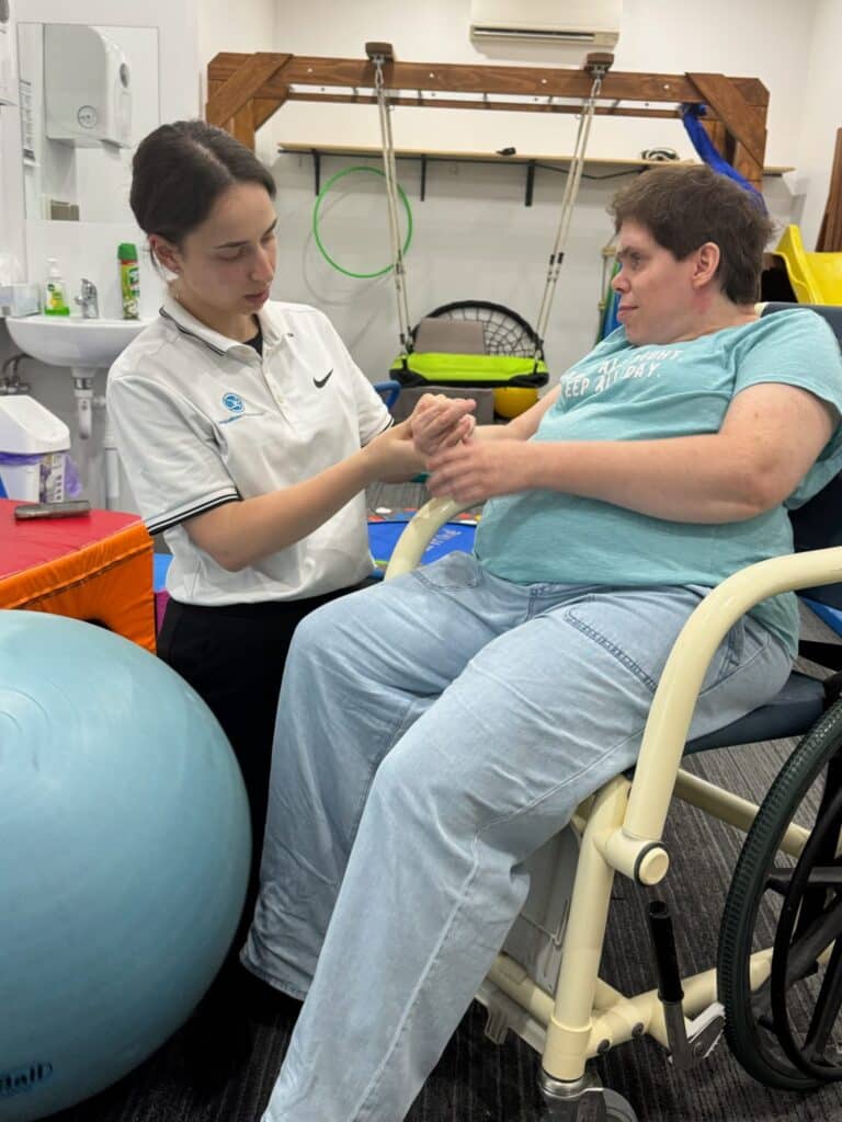 A physical therapist assists a woman in a wheelchair with hand exercises, celebrating each small achievement in a therapy room equipped with rehab equipment.