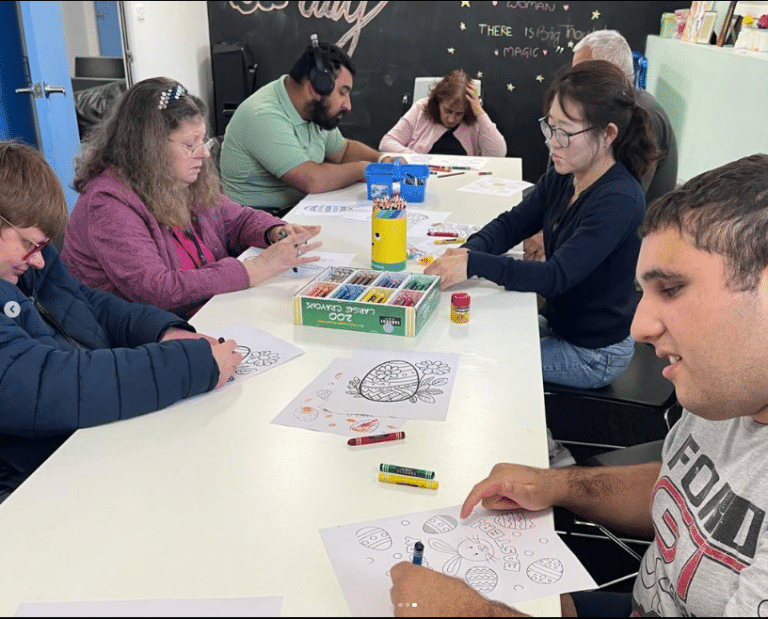 People sitting around a table engage in an Easter craft coloring activity with printed sheets, markers, and crayons. The wall behind them features a chalkboard adorned with motivational writing.