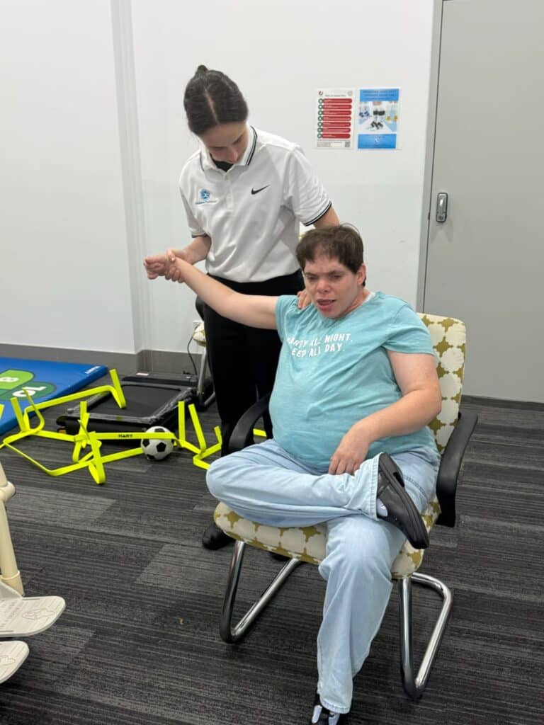 A support worker assists a seated person with mobility exercises in a therapy room equipped with sports and exercise equipment, fostering a sense of achievement with each milestone reached.