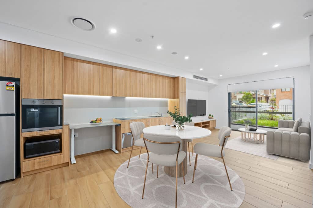 Modern open-plan kitchen and dining area with wooden cabinetry, round dining table with four chairs, and a living space featuring a sofa and large window.
