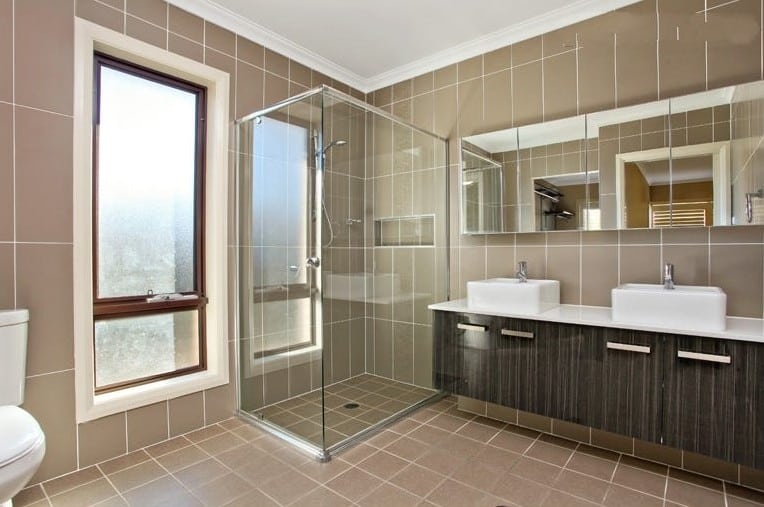 Modern bathroom with glass-enclosed shower, double vanity with vessel sinks, large wall mirror, beige tile walls and floor, and a frosted window.