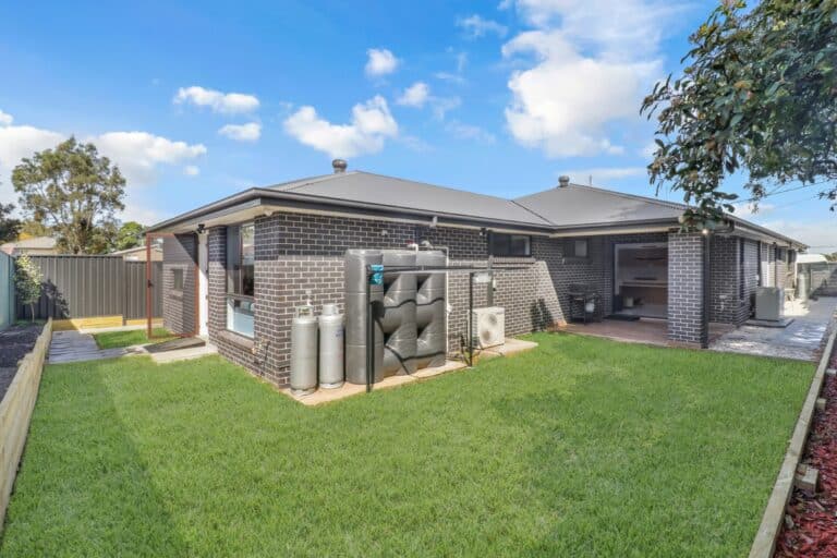 Single-story brick house with a well-maintained backyard, gas bottles, water tank, air conditioning unit, and a covered patio area under a partly cloudy sky.