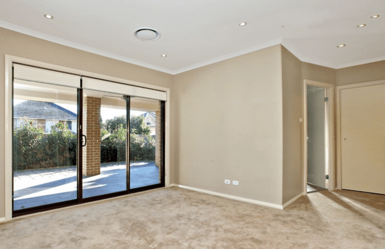 Empty beige-carpeted room with sliding glass doors leading to a covered patio; walls and ceiling are light-colored with recessed lighting.