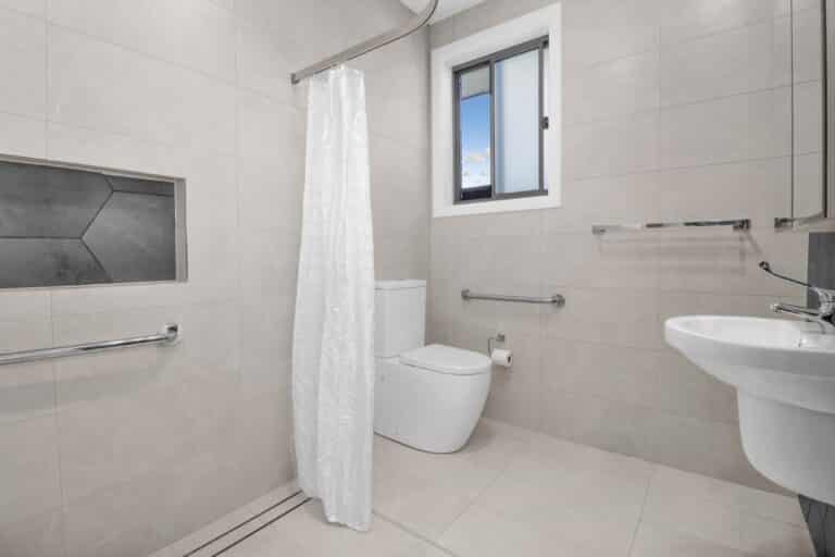 Accessible bathroom with a toilet, wall-mounted sink, shower area with a white curtain, grab bars, built-in shower shelf, and a window.