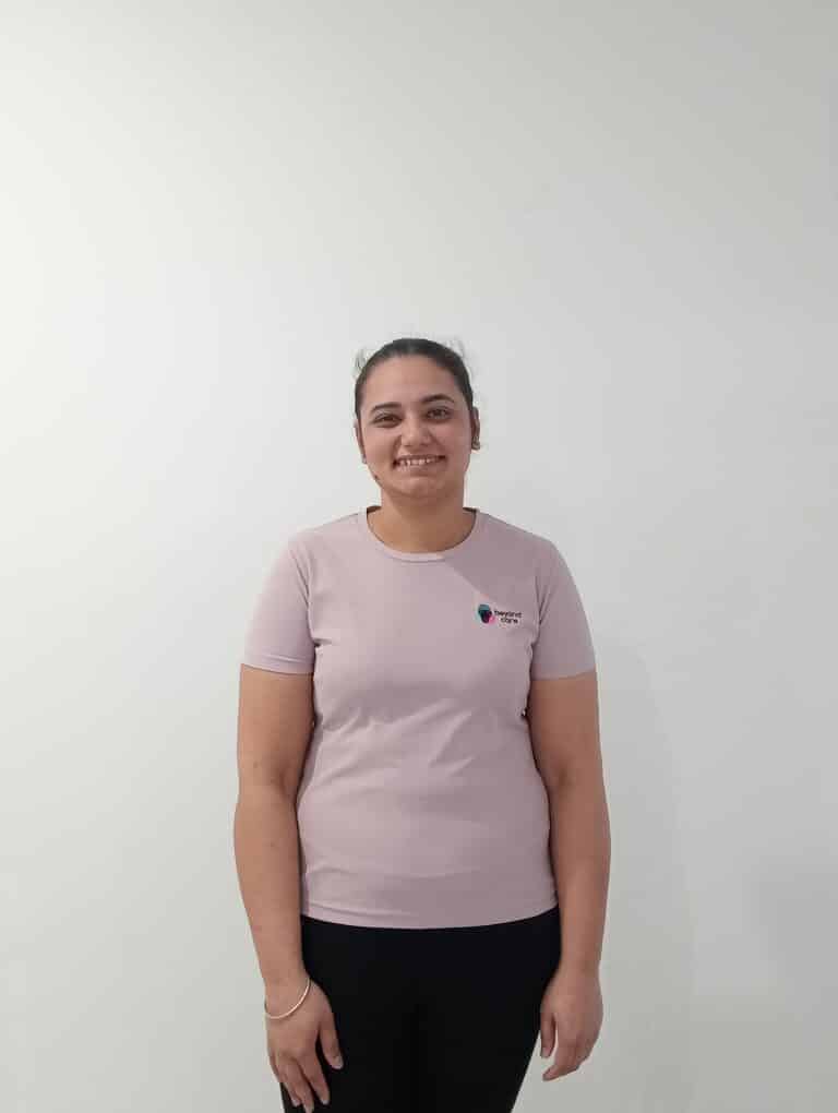 A person with tied-back hair is standing against a plain white wall, wearing a light pink t-shirt and black pants, and smiling at the camera.