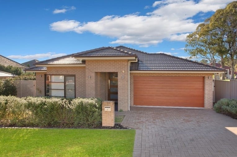 Single-story brick house with a dark tiled roof, double garage with wooden door, paved driveway, front lawn, and a small garden.
