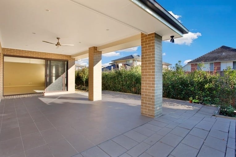 Covered patio area with tiled flooring, brick columns, ceiling fan, and view of backyard with hedges and neighboring houses.