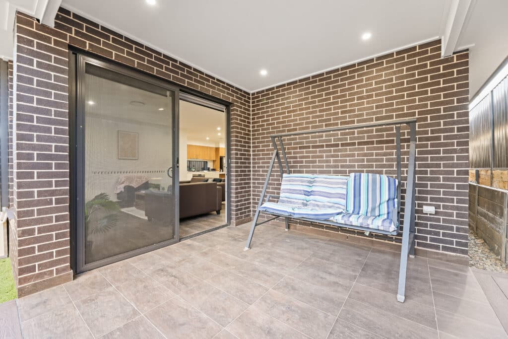 A covered patio with brown brick walls, a tiled floor, a metal-framed swing with striped cushions, and a sliding glass door leading to an indoor living area.