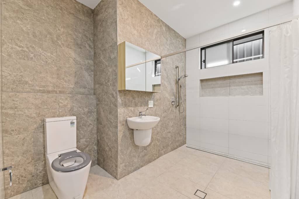 Modern accessible bathroom with a wall-mounted sink, toilet, walk-in shower with grab bar, and large mirror, featuring beige stone tiles and white walls.