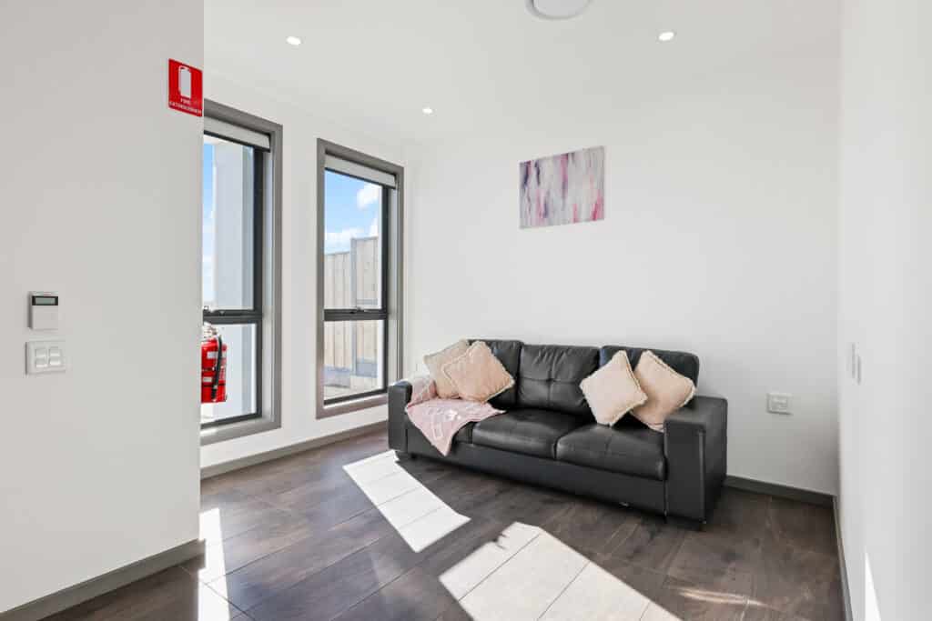 Small, bright room with two tall windows, a black leather sofa with pillows and a pink throw, wall art above, and a fire extinguisher mounted near the windows.