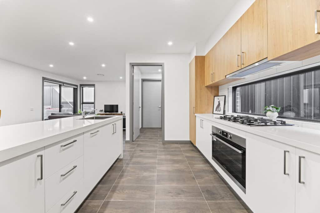 Modern kitchen with white cabinetry, wood upper cabinets, built-in oven, gas stove, large island, and tile flooring, leading to an open living area with windows.