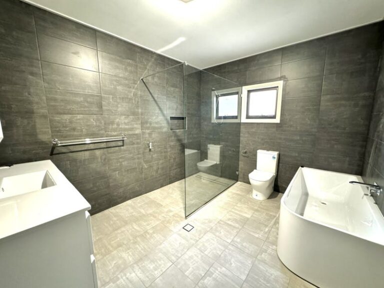 Modern bathroom with gray tiled walls, featuring a walk-in shower, freestanding bathtub, toilet, double sink vanity, and two small windows.