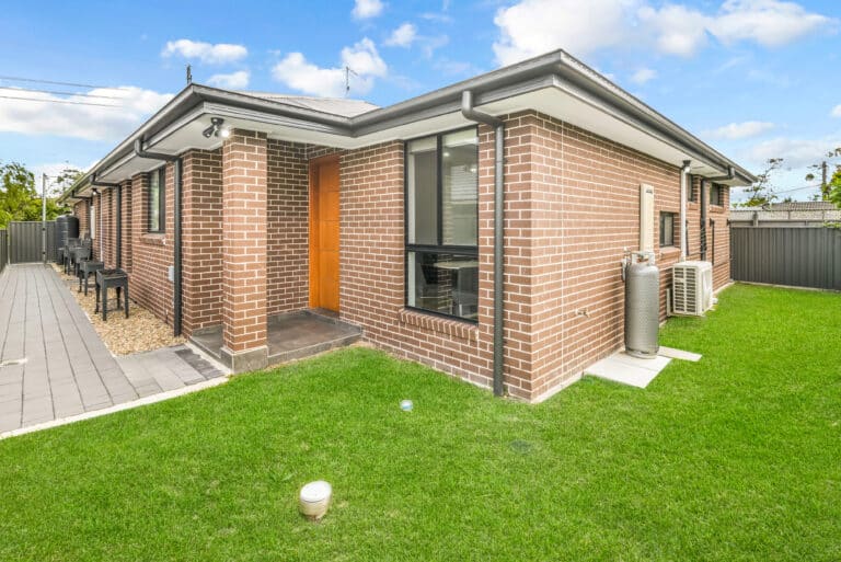 Single-story brick house with a brown door, paved walkway, green lawn, and outdoor air conditioning unit—ideal SIL Accommodation in Sydney with comfort and convenience under a cloudy blue sky.