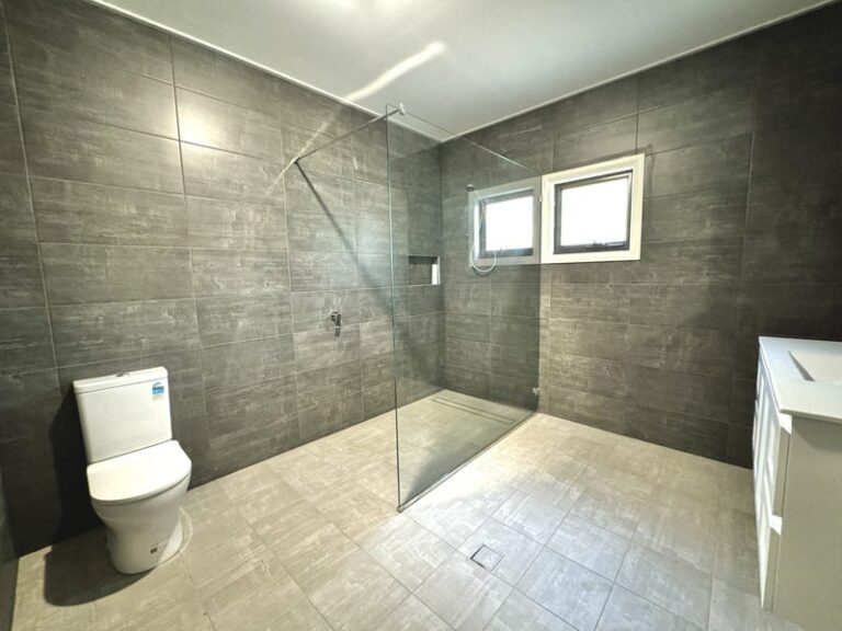 Modern bathroom with gray tiled walls and floor, open walk-in shower with glass partition, white toilet, two windows, and a white vanity unit.
