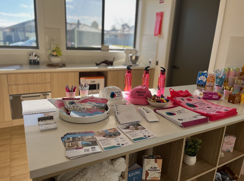 A kitchen counter displays promotional brochures, pink water bottles, hats, tote bags, candy, and various office supplies in a well-lit room.