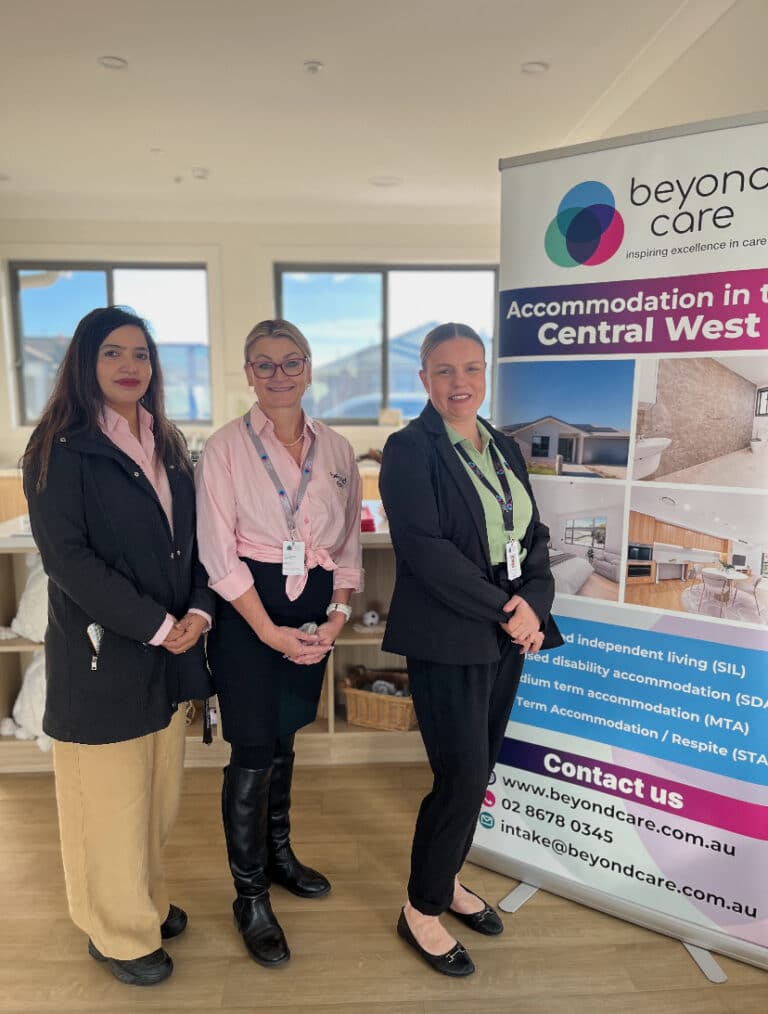Three women stand indoors next to a Beyond Care promotional banner about accommodation services in Central West NSW.