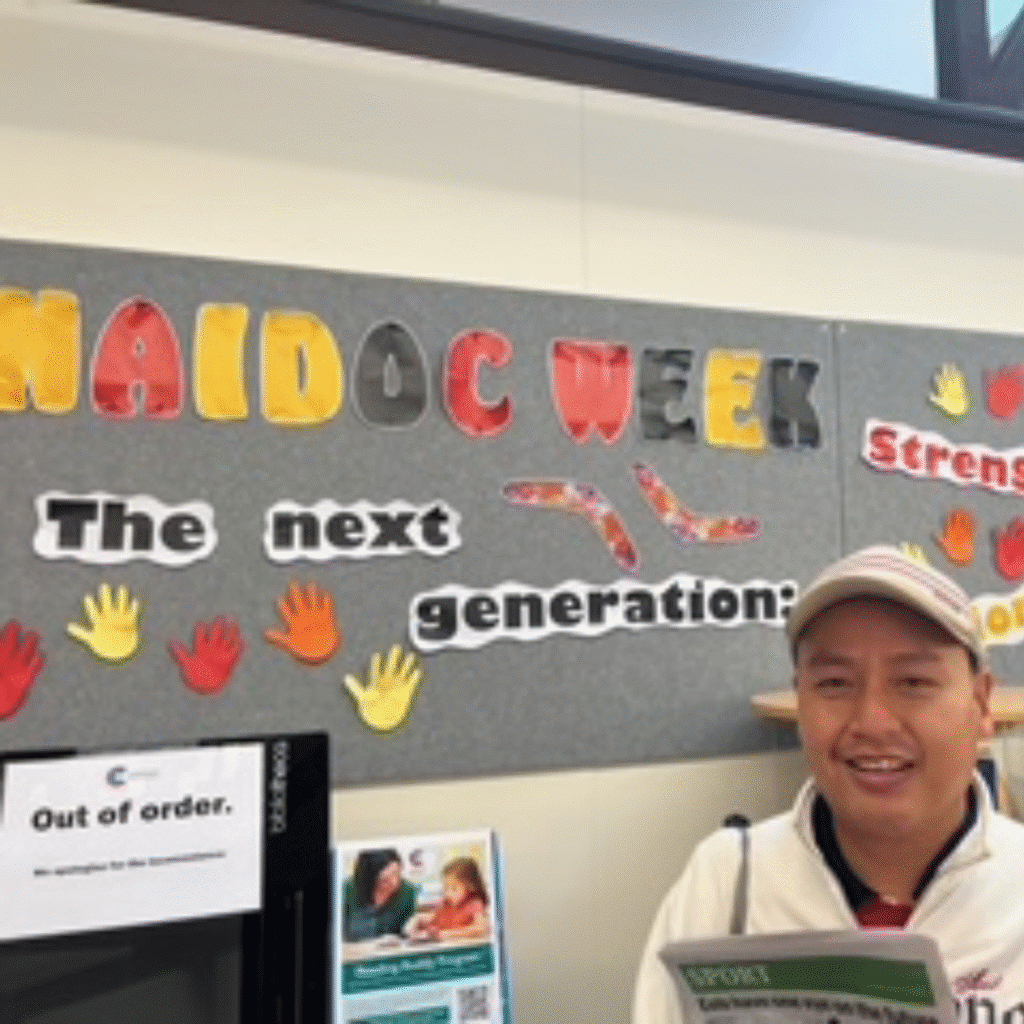 A person stands in front of a bulletin board decorated for NAIDOC Week with colorful letters, handprints, and a sign reading "Out of order.