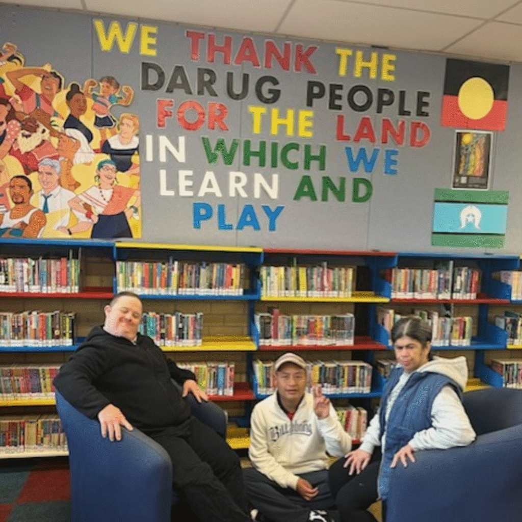 Three people sit in chairs in front of bookshelves and a wall display thanking the Darug people for the land, with colorful text and Aboriginal and Torres Strait Islander flags.