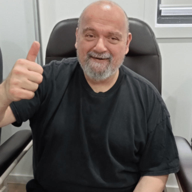 A man with a beard and a black t-shirt sits on a chair, smiling and giving a thumbs up with his right hand.