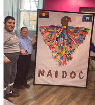 Two people stand beside a large poster featuring a bird made from colorful feathers and the word "NAIDOC," with Aboriginal and Torres Strait Islander flags at the top corners.
