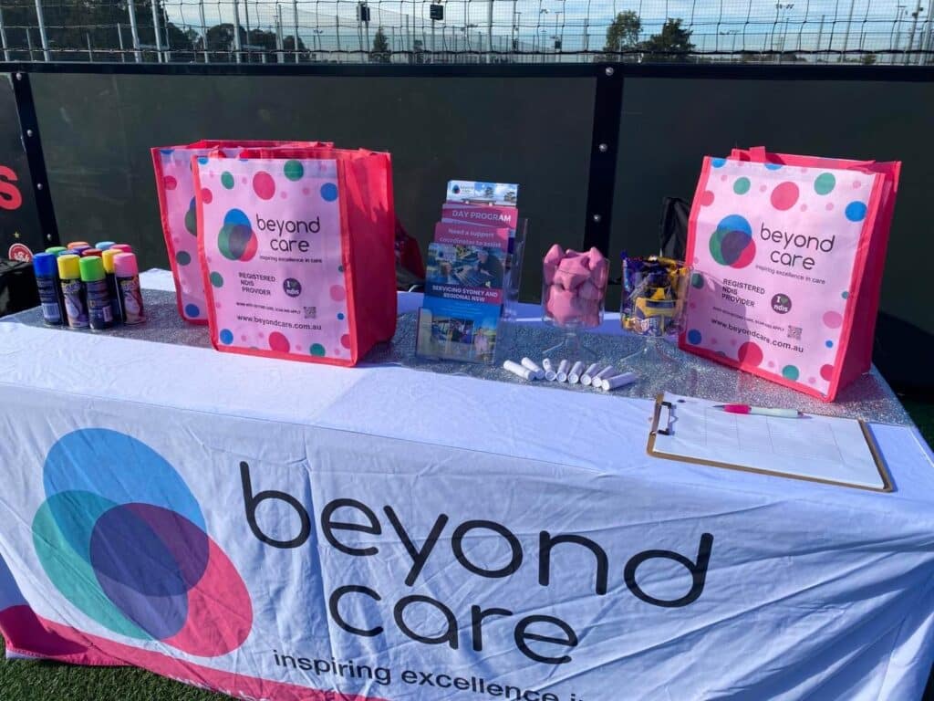 A table covered with a “beyond care” cloth displays pink gift bags, brochures, spray paint cans, and a clipboard set up for a disability-friendly community event at an outdoor gathering.