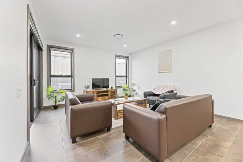 Modern living room with brown sofas, a small TV on a wooden stand, a coffee table, potted plants, and minimal wall decor; large windows provide natural light.