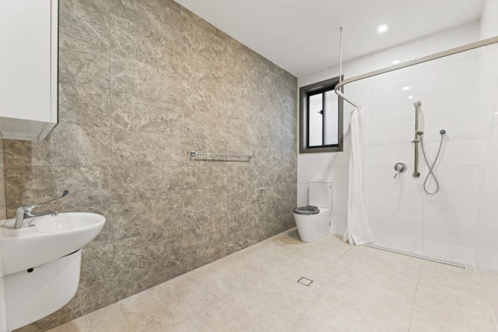 Accessible bathroom with a wall-mounted sink, toilet, and open shower area featuring grab bars, a handheld showerhead, and a curtain. Stone accent wall and beige floor tiles.