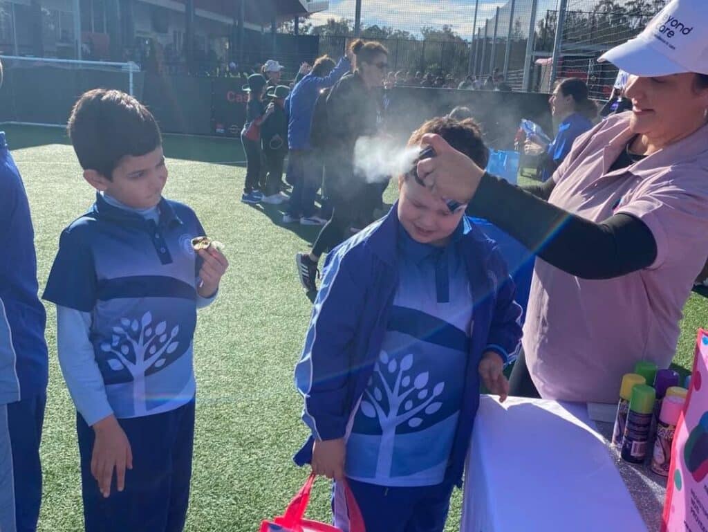 A woman sprays a child's hair while another, holding a medal, stands nearby; both kids wear matching blue uniforms with a tree design at the All Abilities Gala Day, celebrating disability-friendly community events.