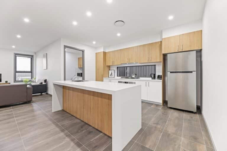 Modern kitchen with wooden cabinets, white countertops, stainless steel appliances, and an island, adjacent to a living area with neutral-toned flooring and walls.