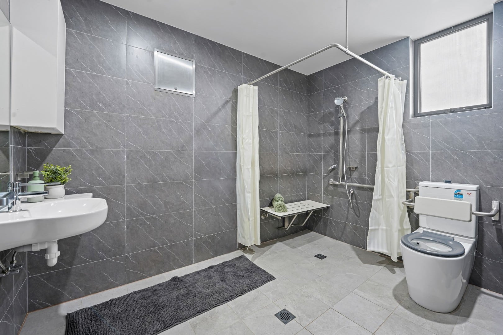 Accessible bathroom within specialist disability accommodation (SDA NDIS), featuring gray tiled walls, a wall-mounted sink, grab bars, a shower area with a foldable bench, and a toilet with support rails.