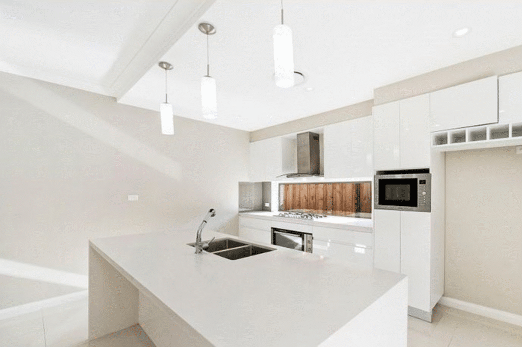Modern white kitchen with an island, built-in appliances, pendant lights, and a wooden backsplash.