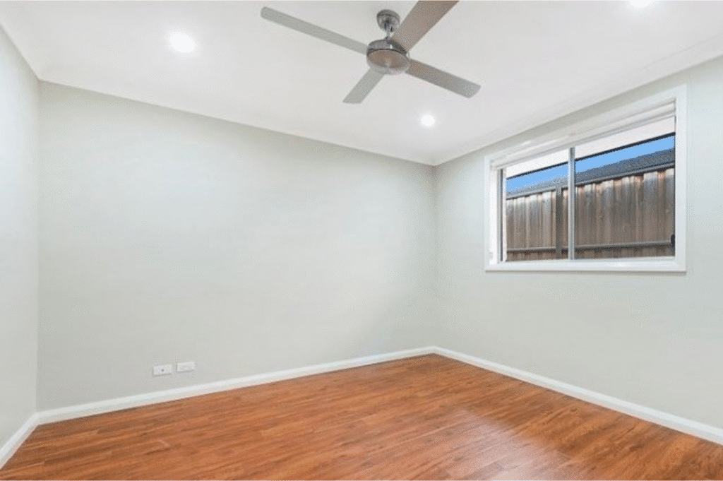 Empty room with white walls, wooden floor, ceiling fan, recessed lights, and a window showing a wooden fence outside.