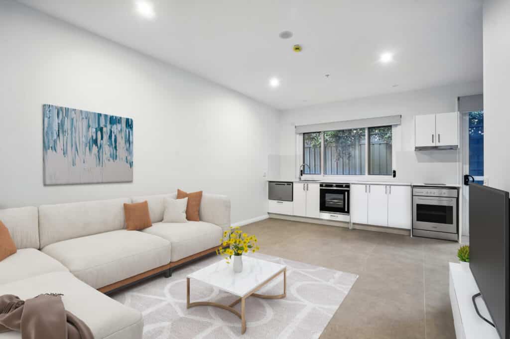 Modern open-plan living room and kitchen designed for Specialist Disability Accommodation (SDA NDIS), featuring white walls, a beige sectional sofa, a coffee table, and accessible built-in appliances along the back wall.