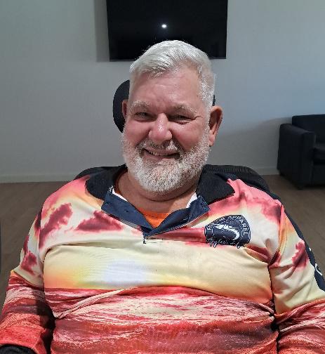 An older man with white hair and a beard is sitting indoors, smiling. He wears a colorful shirt with a landscape print and logo—an inspiring SDA participant story reflecting NDIS success in Beyond Care SDA homes.