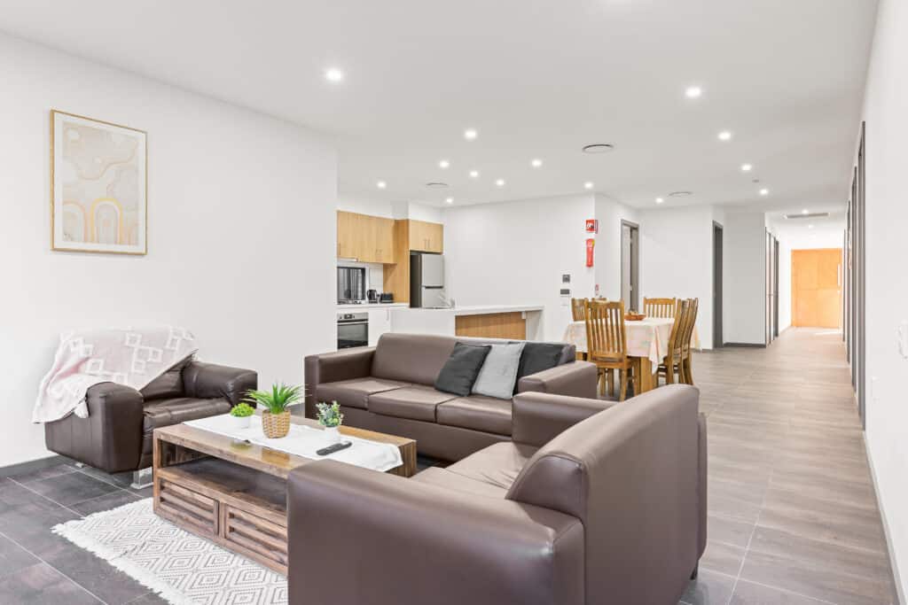 Modern open-plan living room and kitchen featuring brown leather sofas, a dining table, and neutral decor. Bright lighting and a minimalistic style throughout make the space welcoming for those seeking accessible living with NDIS services.