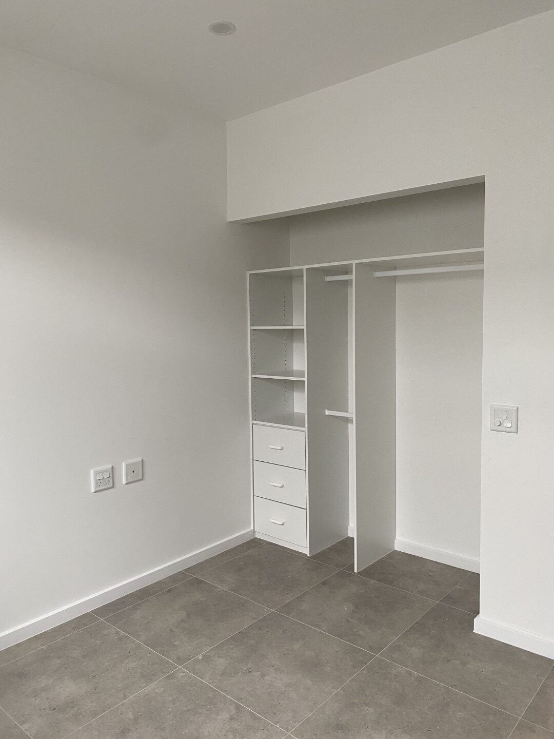 Empty white closet with open shelving, drawers, and hanging rods, set against a plain white wall with a tiled floor.