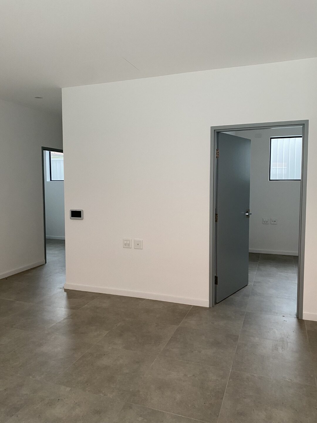 A plain, empty room with light-colored tile flooring, white walls, and two open gray doors leading to adjacent rooms, each with a small window.
