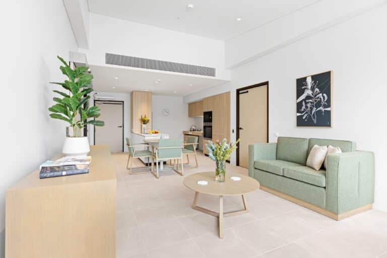 A modern, minimalistic living and dining area with light wood furniture, green sofa, plants, and a wall-mounted artwork in a bright, neutral-toned room.