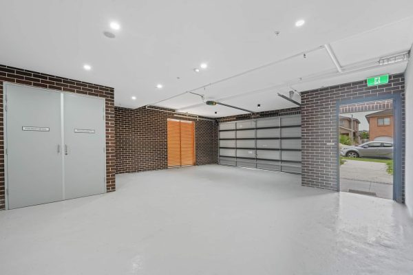 Empty garage with white walls, brick accents, gray double doors, and a large open garage door leading outside. Ceiling lights illuminate the space. Exit sign above one door.