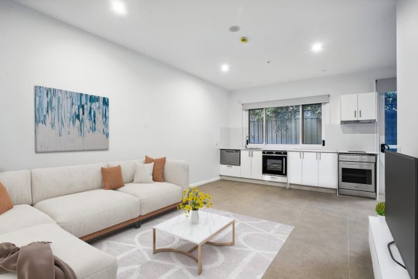 SDA in Penrith 3 Modern open-plan living room and kitchen with white walls, light-colored sofa, coffee table, and large window above the kitchen counter.