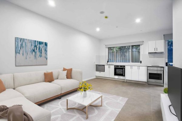 Modern open-plan living room and kitchen with white walls, light-colored sofa, coffee table, and large window above the kitchen counter.