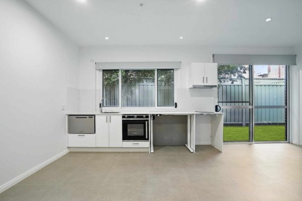 Modern kitchen with white cabinets, built-in oven, and large window looking out to a fenced yard; tiled floor and sliding glass door to the right.