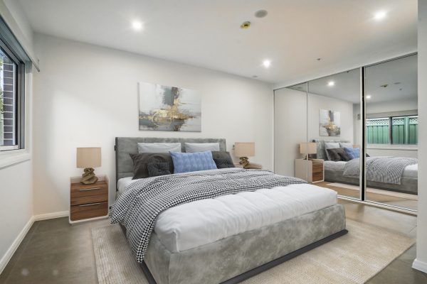 SDA in Penrith 5 Modern bedroom with a gray upholstered bed, two wooden nightstands, abstract wall art, and mirrored sliding closet doors. A window and recessed lighting brighten the room.