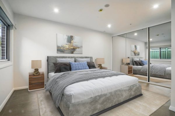 Modern bedroom with a gray upholstered bed, two wooden nightstands, abstract wall art, and mirrored sliding closet doors. A window and recessed lighting brighten the room.