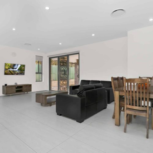 A modern living room with a black leather sofa, wooden coffee and dining tables, a wall-mounted TV, and large sliding doors leading to a patio.