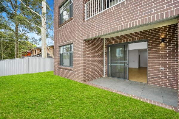 Merrylands SDA 12 Exterior view of a brick building with a patio and sliding glass doors leading to a grassy yard, surrounded by a metal fence.